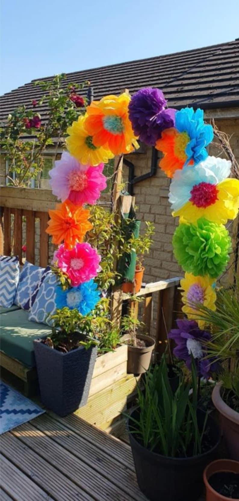 May include: A decorative archway constructed from large, multi-coloured tissue paper flowers in shades of yellow, orange, pink, purple, blue, and green, arranged in a curved formation and positioned outdoors on a wooden deck.