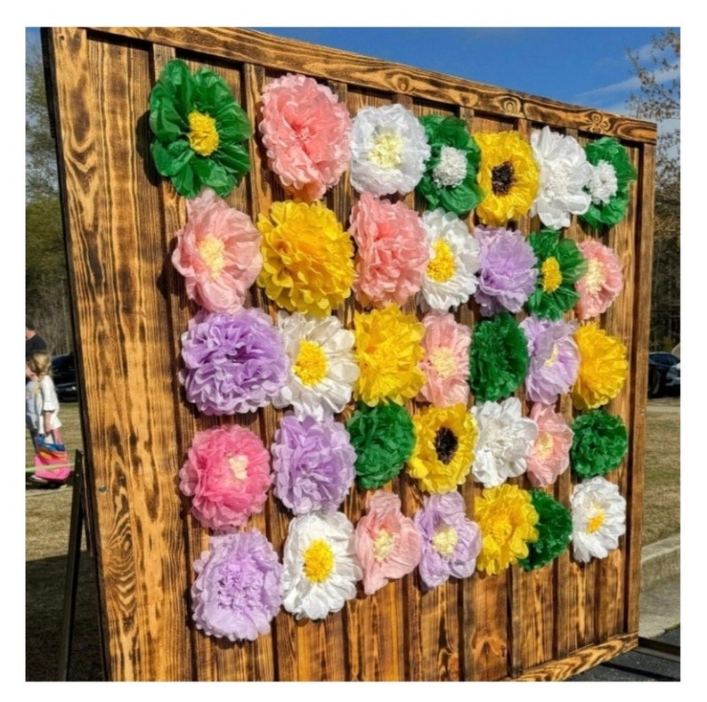 May include: A wooden backdrop decorated with tissue paper flowers in various colors, including pink, yellow, green, white, and purple. The flowers are arranged in a random pattern, creating a festive and colorful display.
