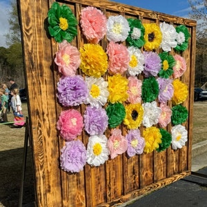 May include: A rustic wooden backdrop decorated with colorful tissue paper flowers in shades of pink, yellow, white, green, and purple. The flowers are arranged in a grid pattern and create a festive and cheerful backdrop.