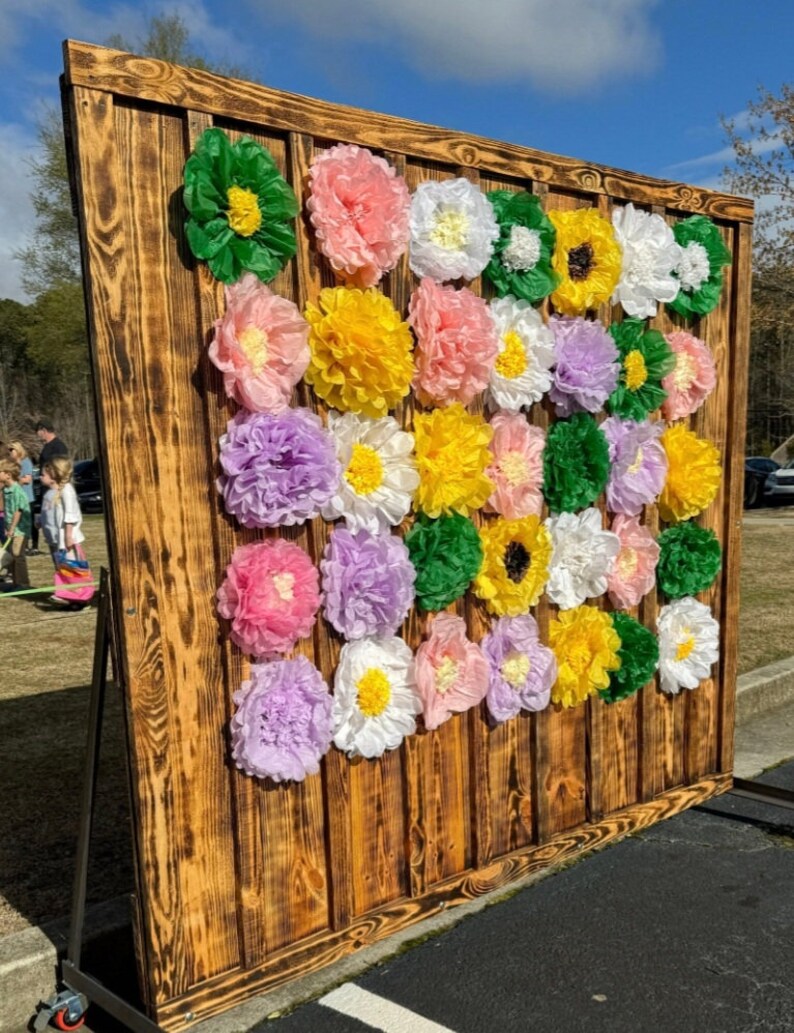 May include: A rustic wooden backdrop decorated with colorful tissue paper flowers in shades of pink, yellow, white, green, and purple. The flowers are arranged in a grid pattern and create a festive and cheerful backdrop.