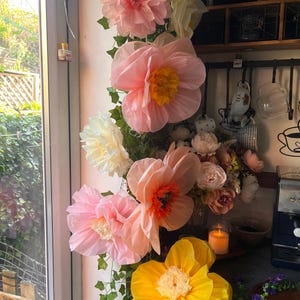 May include: A vertical arrangement of large, layered paper flowers in shades of pink, peach, cream, and yellow, interspersed with green faux ivy vines. The flowers feature ruffled petals and contrasting centres, creating a voluminous display.
