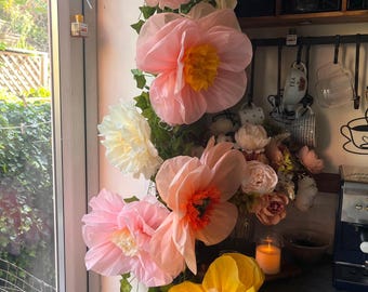 Puede incluir: Un arreglo vertical de grandes flores de papel en capas en tonos rosa, melocotón, crema y amarillo, intercaladas con enredaderas de hiedra artificial verde. Las flores presentan pétalos con volantes y centros en contraste.