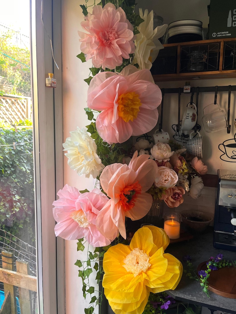May include: A vertical arrangement of large, layered paper flowers in shades of pink, peach, cream, and yellow, interspersed with green faux ivy vines. The flowers feature ruffled petals and contrasting centres, creating a voluminous display.