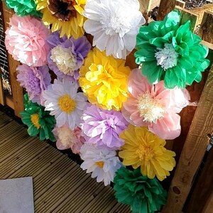 May include: A wall decorated with tissue paper flowers in various colors, including green, yellow, white, pink, and purple. The flowers are arranged in a cascading pattern, creating a festive and whimsical backdrop.