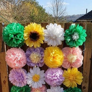May include: A wall decorated with 15 tissue paper flowers in various colors and styles, including green, yellow, white, pink, and purple. The flowers are arranged in a grid pattern on a wooden fence.