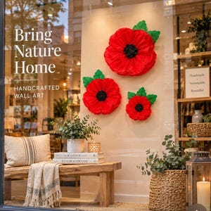 May include: Three large, vibrant red poppy wall art flowers with black centres and green leaves are mounted on a light-coloured wall. The display includes a wooden bench with a striped throw blanket, a potted plant, and a woven basket with greenery.