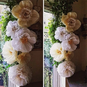 May include: A vertical arrangement of large, layered paper flowers in shades of cream, white, and pale yellow. The flowers feature ruffled, delicate petals with textured centres, creating a voluminous, multi-layered appearance in a floral display.