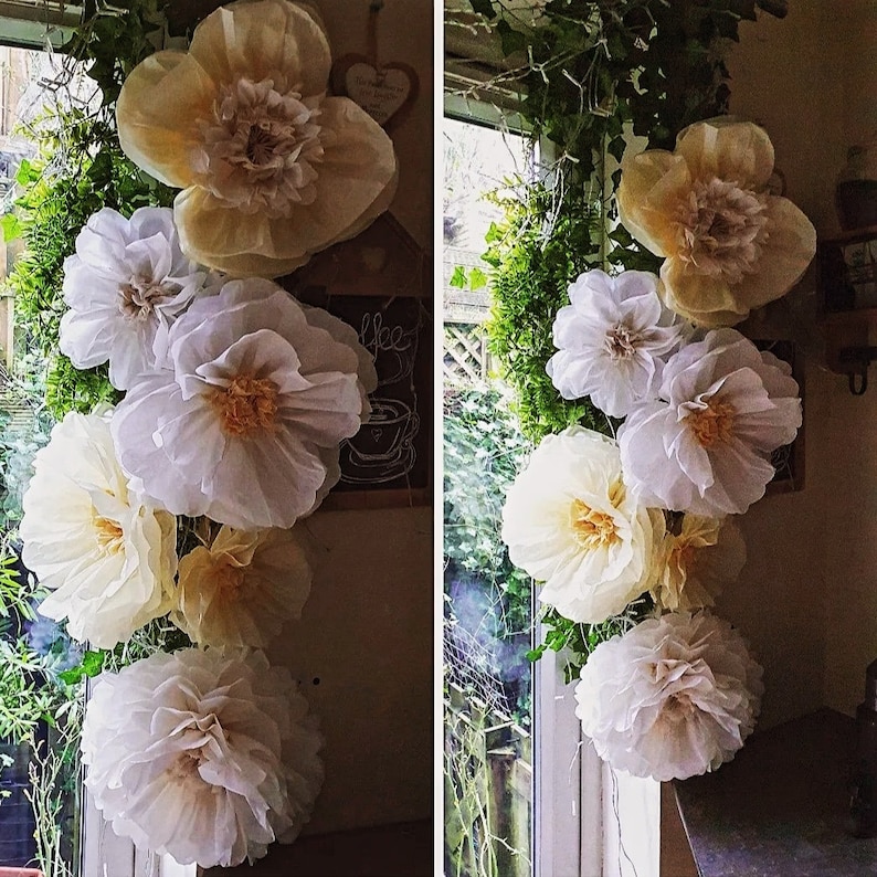 May include: A vertical arrangement of large, layered paper flowers in shades of cream, white, and pale yellow. The flowers feature ruffled, delicate petals with textured centres, creating a voluminous, multi-layered appearance in a floral display.