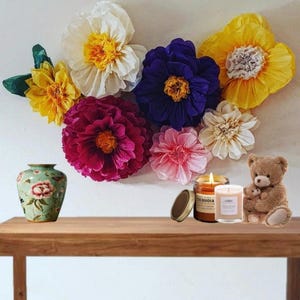 May include: A collection of large, colourful paper flowers in shades of white, yellow, purple, magenta, and pink arranged on a wall. Below, a light green floral vase, two glass candles with labels reading Sequoia and a small brown teddy bear.