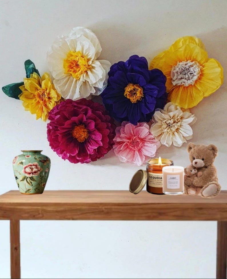 May include: A collection of large, colourful paper flowers in shades of white, yellow, purple, magenta, and pink arranged on a wall. Below, a light green floral vase, two glass candles with labels reading Sequoia and a small brown teddy bear.