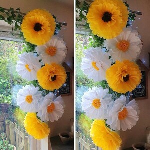May include: A vertical arrangement of large, layered paper flowers in bright yellow and white. The flowers feature ruffled petals and dark or yellow centres, interspersed with green faux ivy vines, creating a decorative floral garland display.