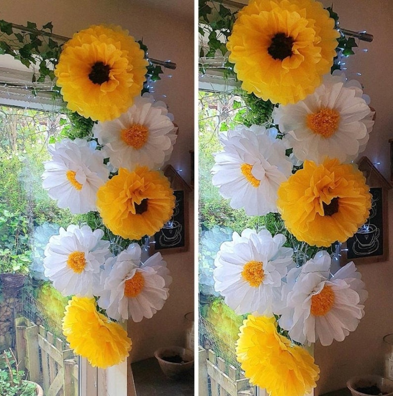 May include: A vertical arrangement of large, layered paper flowers in bright yellow and white. The flowers feature ruffled petals and dark or yellow centres, interspersed with green faux ivy vines, creating a decorative floral garland display.