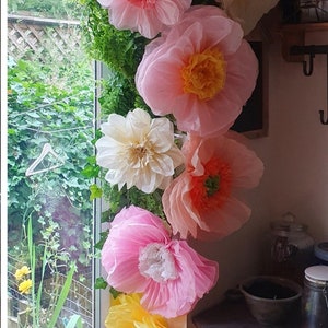 May include: A vertical arrangement of large, layered paper flowers in shades of pink, cream, and yellow, interspersed with green artificial foliage. The flowers feature ruffled petals and textured centres, creating a decorative floral display.