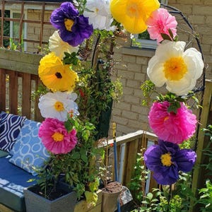 May include: A garden arch decorated with large, vibrant paper flowers in shades of yellow, white, pink and purple. The arch is positioned over a wooden planter box containing green climbing plants, with additional potted plants nearby.
