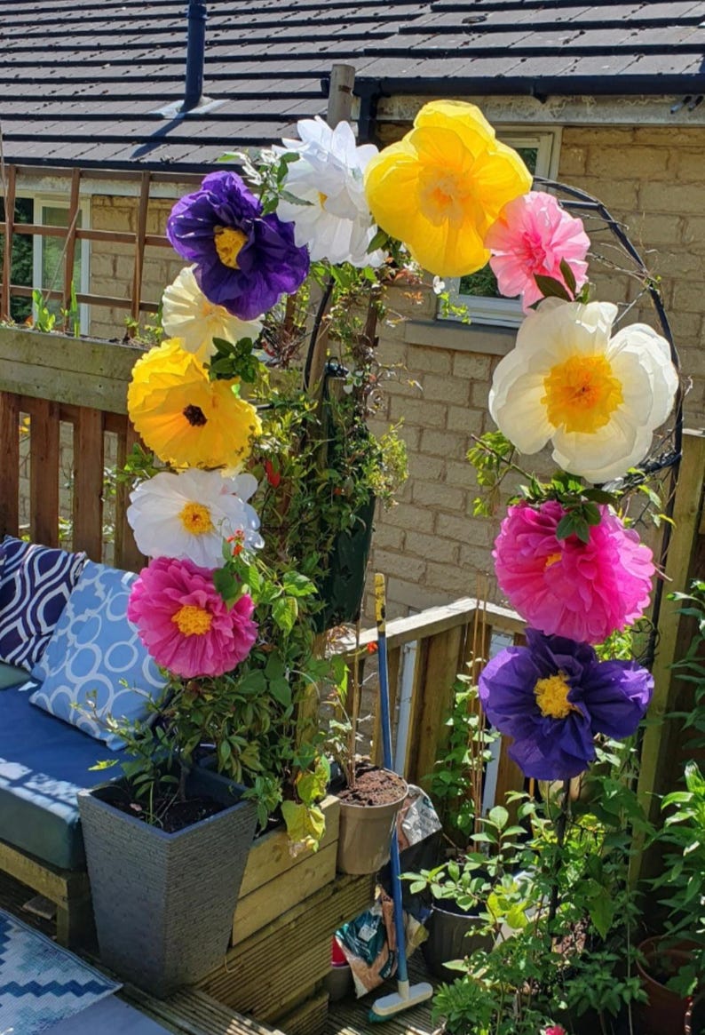 May include: A garden arch decorated with large, vibrant paper flowers in shades of yellow, white, pink and purple. The arch is positioned over a wooden planter box containing green climbing plants, with additional potted plants nearby.