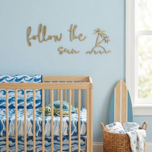 Op de afbeelding: Een kinderkamer met een lichtblauwe muur heeft een houten wieg met blauw-wit golvenpatroon beddengoed. Een houten bord aan de muur leest "follow the sun" met een palmboomontwerp. Een surfplank en een rieten mand met dekens maken het strandthema compleet.