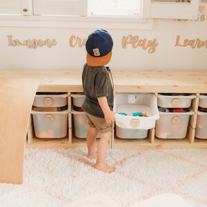 May include: A child wearing a blue baseball cap with the number 5 on it is standing in front of a wooden storage unit with white bins. The bins are labeled with the words "Imagine", "Create", "Play", and "Learn". The child is holding a bin filled with toys. The storage unit is on a white fluffy rug.