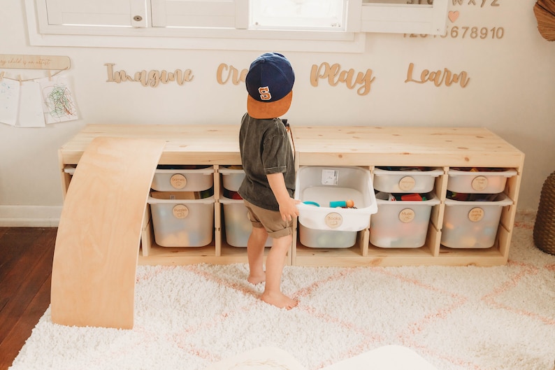 May include: A child wearing a blue baseball cap with the number 5 on it is standing in front of a wooden storage unit with white bins. The bins are labeled with the words "Imagine", "Create", "Play", and "Learn". The child is holding a bin filled with toys. The storage unit is on a white fluffy rug.