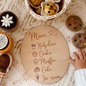 May include: A wooden round sign with the word "Menu" and a heart symbol. The sign lists different food items with corresponding icons: Babychino, Cookie, Muffin, Cake, Ice-cream.