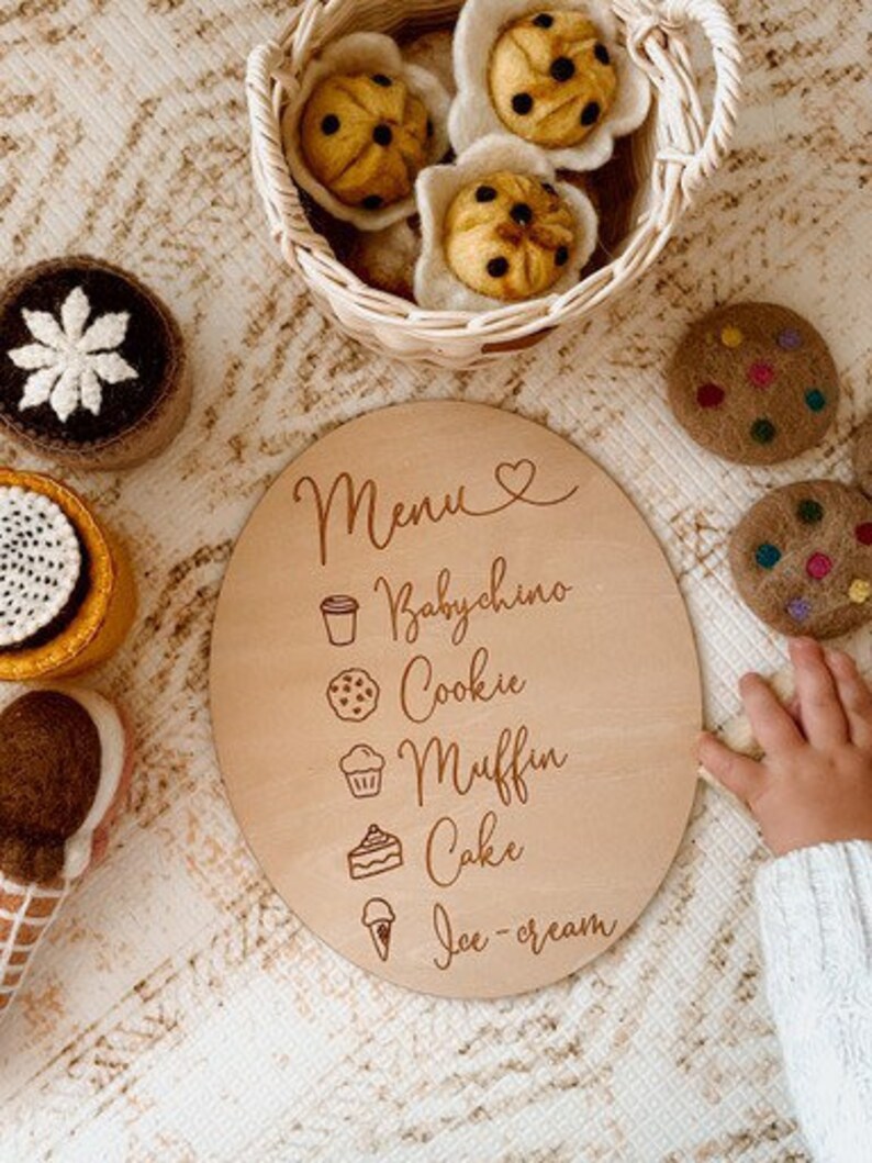 May include: A wooden round sign with the word "Menu" and a heart symbol. The sign lists different food items with corresponding icons: Babychino, Cookie, Muffin, Cake, Ice-cream.