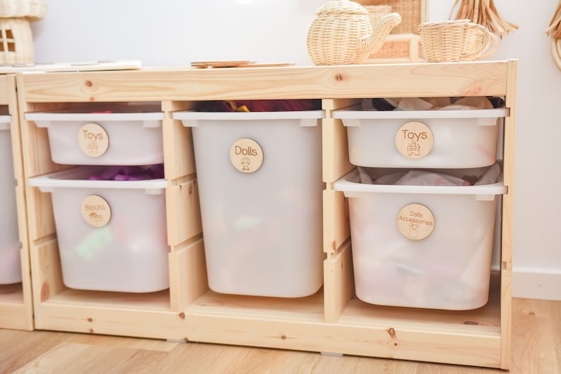 May include: A wooden shelf unit with six white plastic storage bins. Each bin has a wooden circle label with a different category written on it: "Toys", "Dolls", "Books", "Toys", "Doll Accessories", and "Bears".