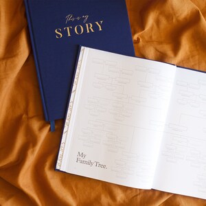 May include: A navy blue hardcover journal with the text "This is my STORY" on the front cover. The journal is open to a page with a blank family tree chart and the text "My Family Tree."