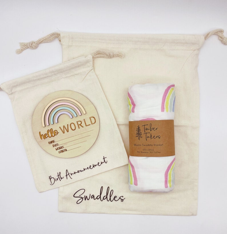 Puede incluir: Una placa de anuncio de nacimiento de madera con forma de arco&iacute;ris con el texto "hello WORLD" y espacios para el nombre, la fecha, el peso y la longitud. La placa est&aacute; en una bolsa de muselina blanca con el texto "Birth Announcement". Una segunda bolsa de muselina blanca con el texto "Swaddles" contiene una manta de muselina de arco&iacute;ris con el texto "Timber Takers Muslin Swaddle Blanket 120 x 120cm 70% Bamboo 30% Cotton".