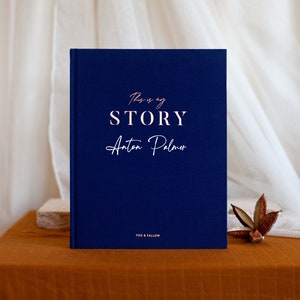 May include: A navy blue hardcover journal with the text "This is my STORY Anton Palmer" in rose gold lettering. The journal is on a brown surface.