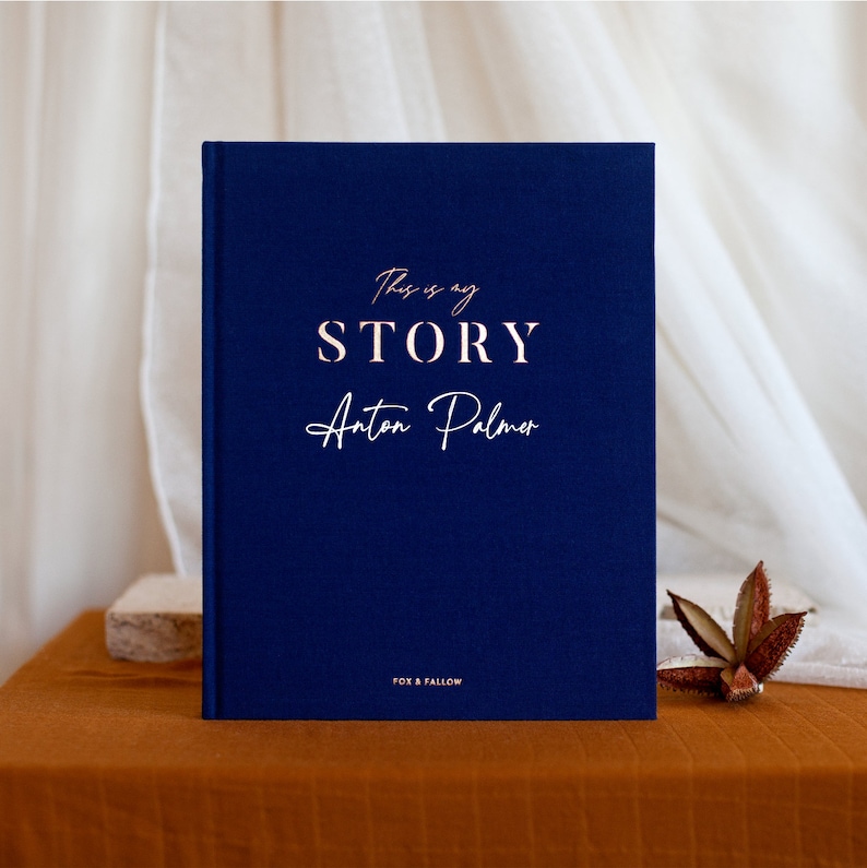 May include: A navy blue hardcover journal with the text "This is my STORY Anton Palmer" in rose gold lettering. The journal is on a brown surface.
