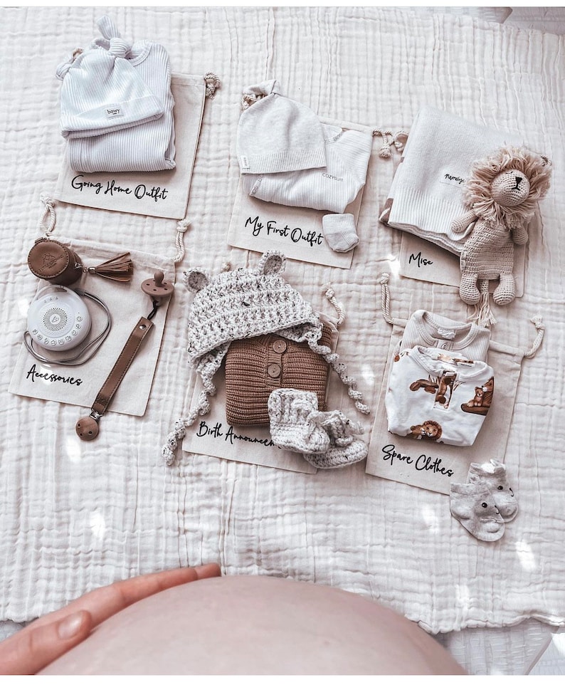Puede incluir: Una colecci&oacute;n de art&iacute;culos para beb&eacute;s organizados en bolsas de tela blanca. Las bolsas est&aacute;n etiquetadas con lo siguiente: "Going Home Outfit", "My First Outfit", "Miss", "Birth Announcement" y "Spare Clothes". Los art&iacute;culos incluyen un body blanco, un gorro blanco, un gorro de punto marr&oacute;n, un jersey de punto marr&oacute;n, un par de botines de punto marr&oacute;n, una manta blanca, un body blanco y marr&oacute;n, un par de calcetines grises, una m&aacute;quina de ruido blanco, un chupete y un le&oacute;n de peluche.