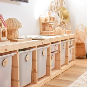 May include: A wooden storage unit with six clear plastic bins. The bins are labeled with wooden tags that say "Cars", "Dinosaurs", "Blocks", "Books", "Puzzles", and "Art".