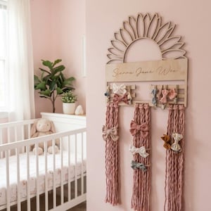 May include: Wooden hair bow organiser in the shape of a sunflower, with the name "Sienna Jane Wei" engraved. It has rows for bows and three pink yarn tassels. Various bows in pink, white, and beige are displayed, creating a charming nursery decor.