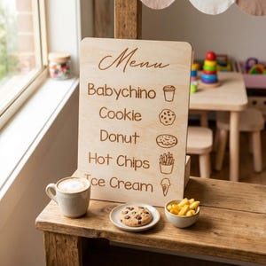 Puede incluir: Pizarra de men&uacute; de madera con texto grabado e ilustraciones de alimentos. El men&uacute; enumera "Babychino", "Cookie", "Donut", "Hot Chips" e "Ice Cream". Una taza de caf&eacute;, una galleta y un cuenco de patatas fritas est&aacute;n sobre la superficie de madera.