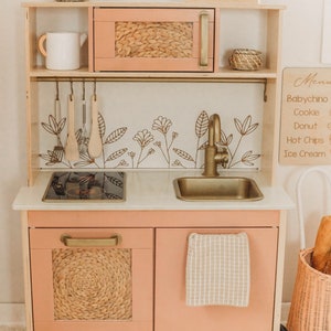 Puede incluir: Una cocina de juguete de madera en color melocot&oacute;n claro con herrajes dorados. Incluye microondas, fregadero y encimera. Los detalles florales decorativos y tejidos a&ntilde;aden encanto. Incluye una pizarra de men&uacute; y una peque&ntilde;a cesta.