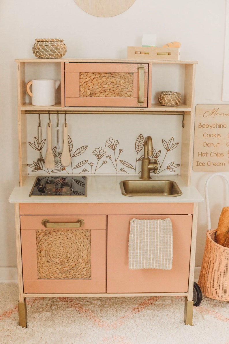 Puede incluir: Una cocina de juguete de madera en color melocot&oacute;n claro con herrajes dorados. Incluye microondas, fregadero y encimera. Los detalles florales decorativos y tejidos a&ntilde;aden encanto. Incluye una pizarra de men&uacute; y una peque&ntilde;a cesta.