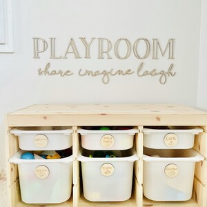 May include: A wooden playroom storage unit with six white plastic bins. Each bin has a wooden label with a different category of toys: Costumes, Arts & Crafts, Cars, Games, Trains, and Puzzles.