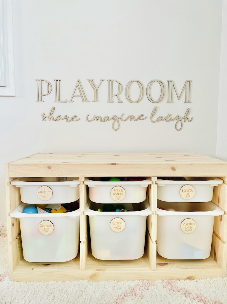 May include: A wooden playroom storage unit with six white plastic bins. Each bin has a wooden label with a different category of toys: Costumes, Arts & Crafts, Cars, Games, Trains, and Puzzles.