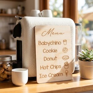 Puede incluir: Un tablero de men&uacute; de madera con la palabra "Men&uacute;" en la parte superior, que enumera "Babychino", "Cookie", "Donut", "Hot Chips" e "Ice Cream". El tablero est&aacute; junto a una cafetera blanca y un tarro de galletas.