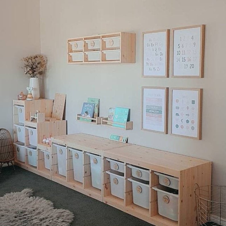 May include: A wooden shelf unit with multiple storage bins, a shelf with books, and a wall with alphabet and number charts. The shelf unit is made of light wood and has a natural finish. The storage bins are white and have labels on them. The alphabet and number charts are printed on white paper and are framed in light wood.