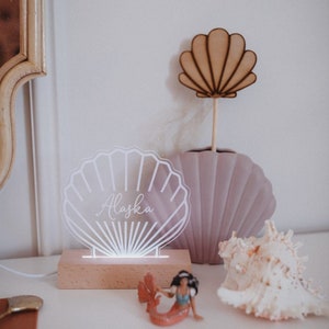 Peut inclure: Une veilleuse en forme de coquillage en acrylique blanc avec le nom "Alaska" inscrit dessus. La lumière est sur une base en bois et est allumée. Il y a un vase en forme de coquillage rose et un coquillage blanc sur la table.