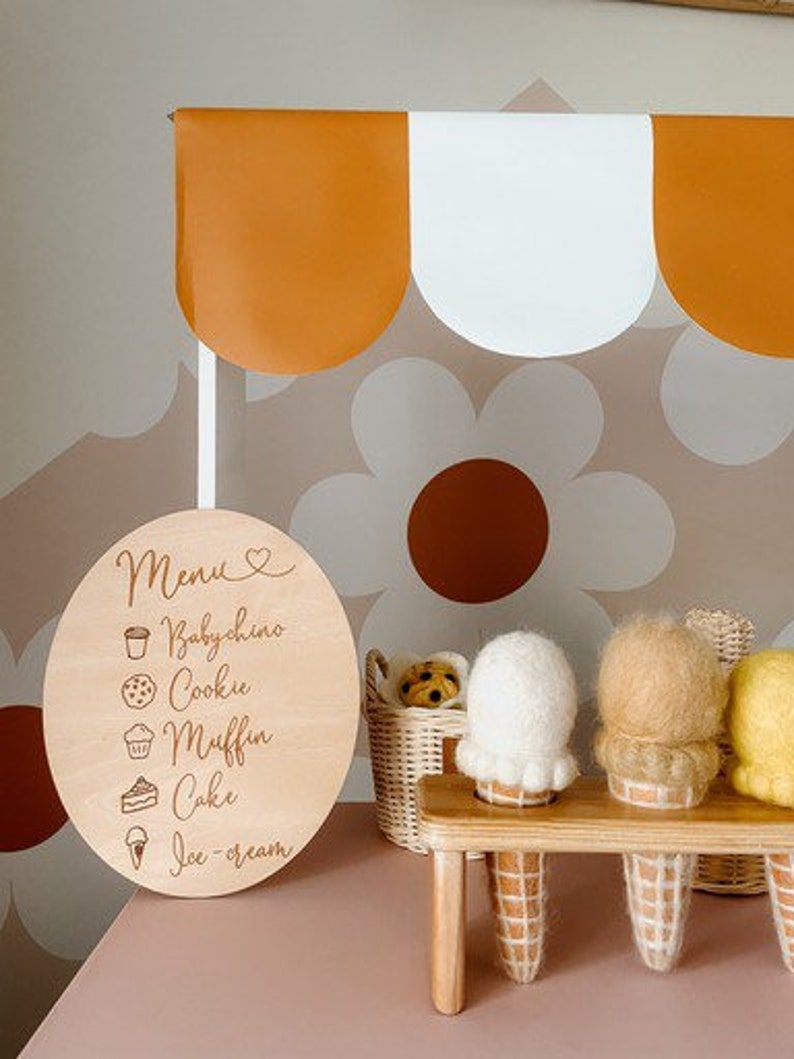 May include: A wooden sign with a menu for a pretend ice cream shop. The menu lists: Babychino, Cookie, Muffin, Cake, and Ice Cream. There are four felt ice cream cones on a wooden stand.