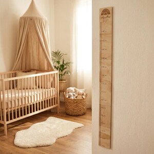 Könnte beinhalten: Ein hölzernes Wachstumsmessgerät mit einem Regenbogen-Design und dem Text "ruby willow" ist an einer Wand befestigt. Die Messlatte hat Markierungen in Zentimetern. Die Kinderzimmereinrichtung umfasst ein Kinderbett, ein Baldachin und einen Weidenkorb mit Stofftieren.