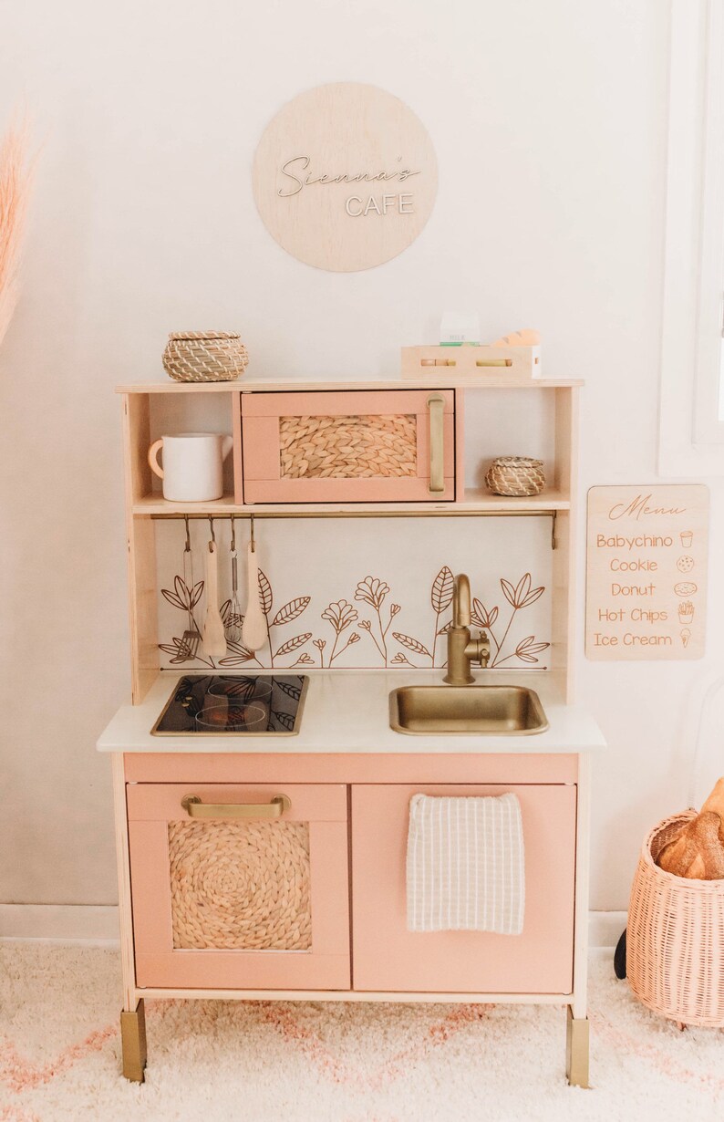 Puede incluir: Una cocina de juguete de madera en rosa suave con herrajes dorados. Cuenta con microondas, fregadero, estufa y almacenamiento. Incluye un letrero que dice "Sienna's Cafe" y una pizarra de men&uacute;. La cocina tiene un dise&ntilde;o floral y una textura tejida en las puertas.