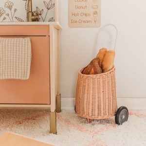 Puede incluir: Una cocina de juguete de madera con un grifo dorado y un men&uacute; con alimentos. Un carrito de mimbre rosa contiene pan. Un letrero de madera con la palabra "curvy" est&aacute; en el suelo.