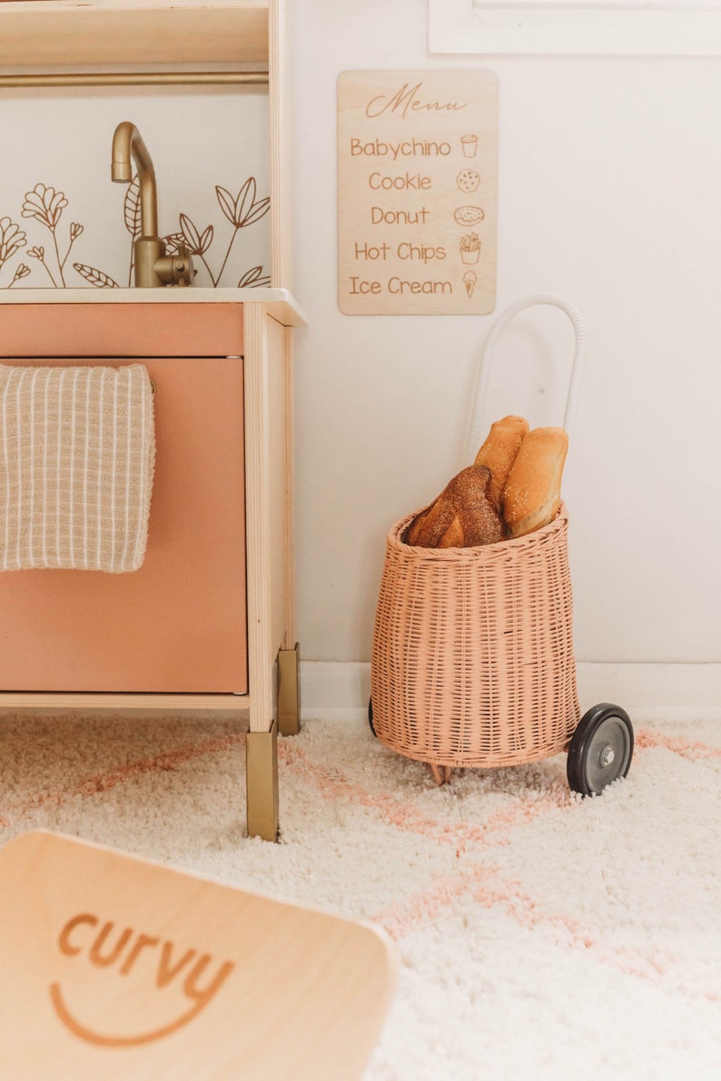 Puede incluir: Una cocina de juguete de madera con un grifo dorado y un men&uacute; con alimentos. Un carrito de mimbre rosa contiene pan. Un letrero de madera con la palabra "curvy" est&aacute; en el suelo.