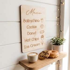 Puede incluir: Un letrero de men&uacute; de madera con la palabra "Men&uacute;" en escritura cursiva. El men&uacute; enumera "Babychino", "Cookie", "Donut", "Hot Chips" y "Ice Cream". Una taza de caf&eacute;, una galleta con chispas de chocolate y una rosquilla est&aacute;n en un estante de madera.
