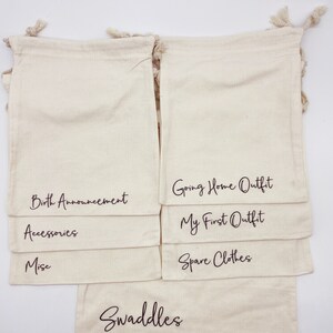 Puede incluir: Conjunto de 10 bolsas de algod&oacute;n blanco con cord&oacute;n de ajuste y texto negro. Las bolsas est&aacute;n etiquetadas: Swaddles, Spare Clothes, My First Outfit, Going Home Outfit, Accessories, Misc, Birth Announcement.