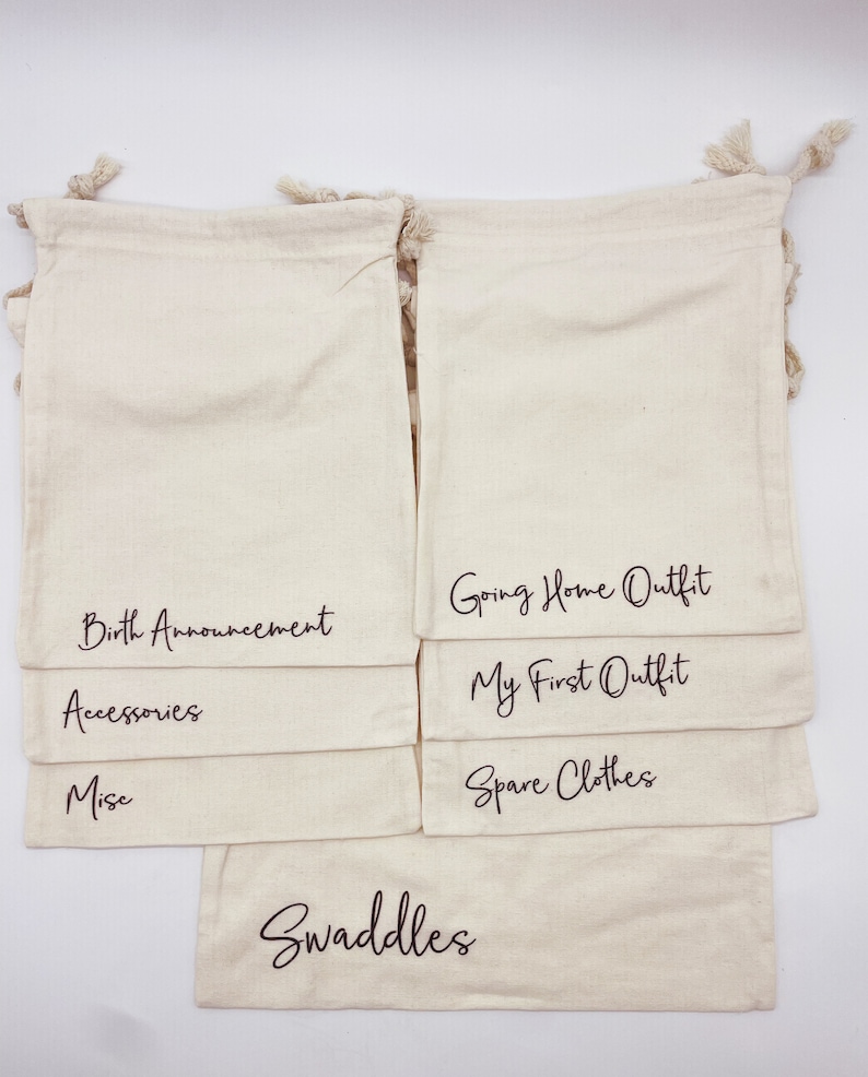 Puede incluir: Conjunto de 10 bolsas de algod&oacute;n blanco con cord&oacute;n de ajuste y texto negro. Las bolsas est&aacute;n etiquetadas: Swaddles, Spare Clothes, My First Outfit, Going Home Outfit, Accessories, Misc, Birth Announcement.