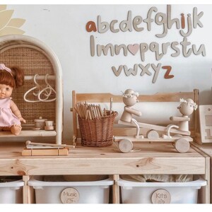 May include: A wooden playset with a seesaw, a dollhouse, and a xylophone. The playset is painted in natural wood tones and features a doll, a basket of pencils, and a set of wooden letters that spell out the alphabet. The letters are painted in a light brown color with a pink heart in the middle of the word "love".