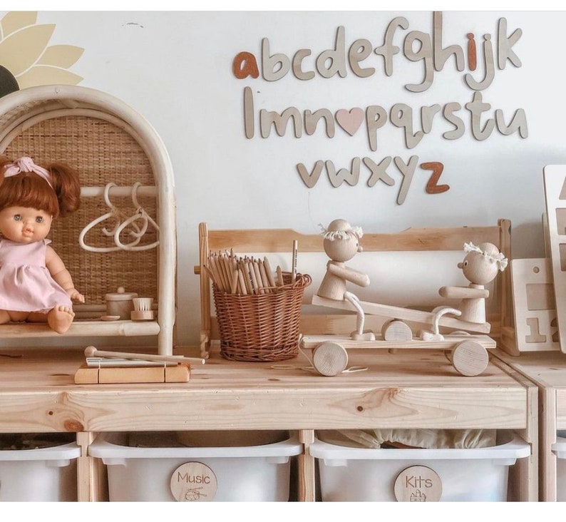 May include: A wooden playset with a seesaw, a dollhouse, and a xylophone. The playset is painted in natural wood tones and features a doll, a basket of pencils, and a set of wooden letters that spell out the alphabet. The letters are painted in a light brown color with a pink heart in the middle of the word "love".